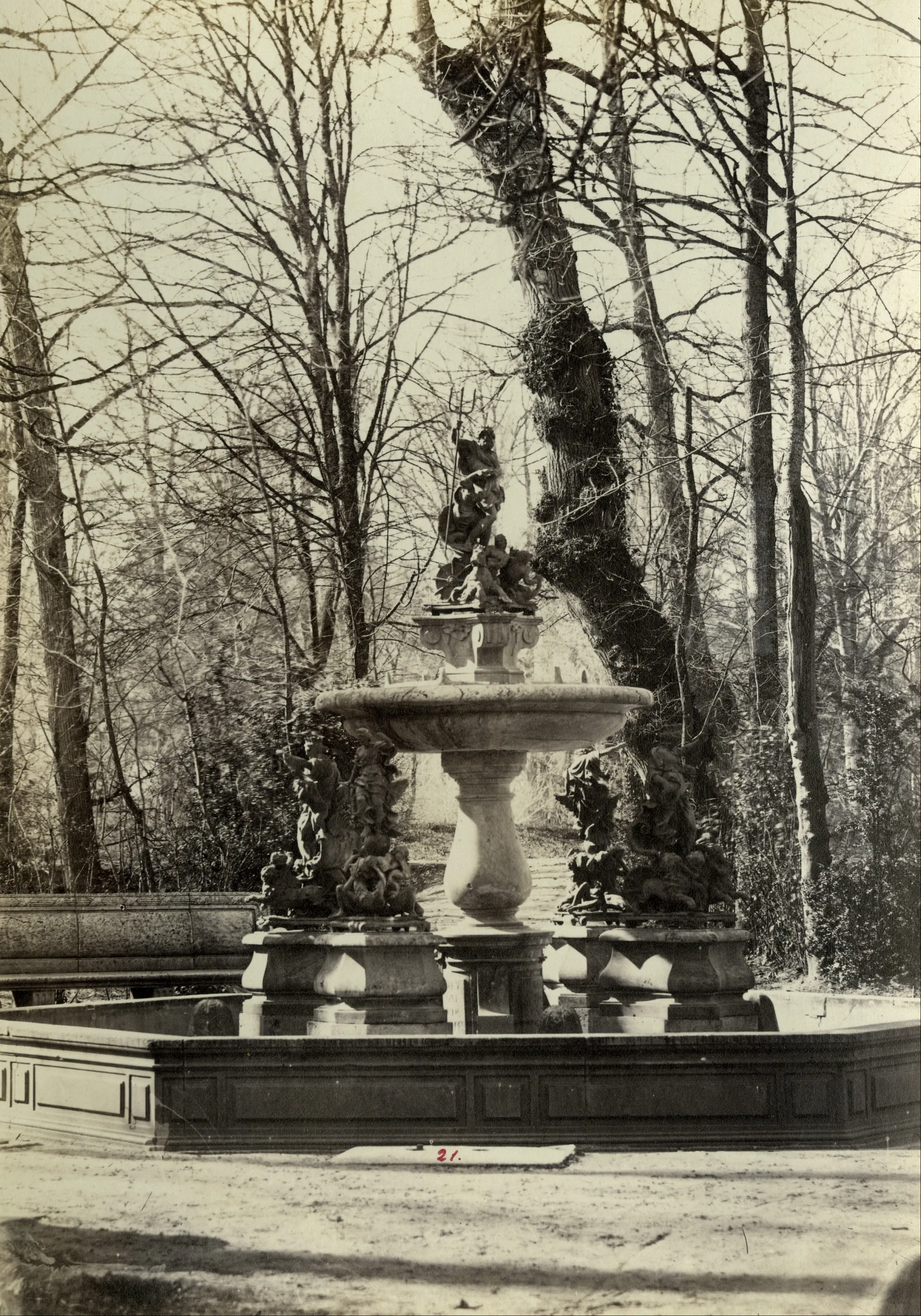 Fotografía antigua de una fuente de mármol de varios cuerpos entre árboles sin hojas.