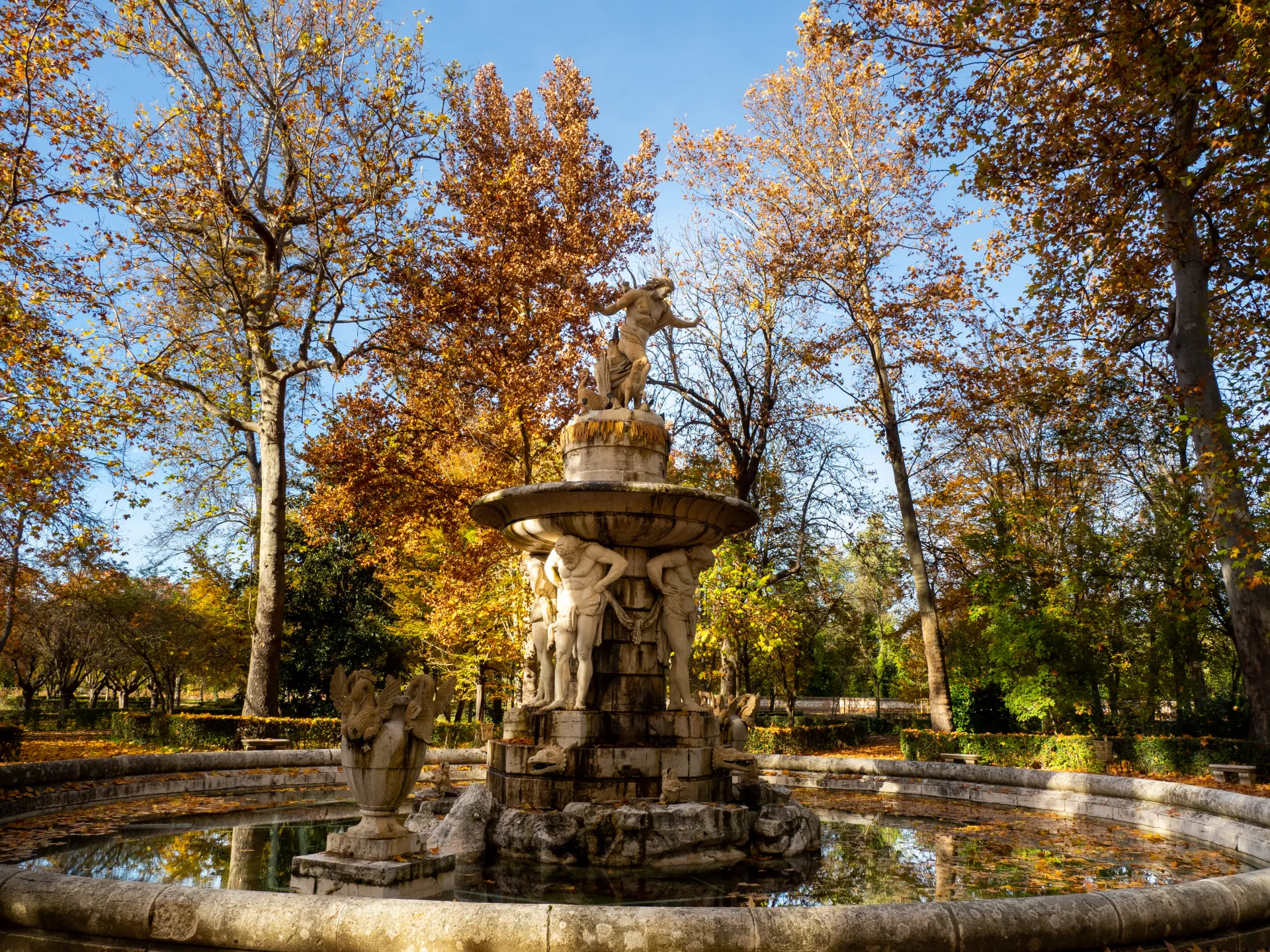 Fotografía de una fuente de piedra de varios cuerpos en el jardín, con arbolado otoñal.