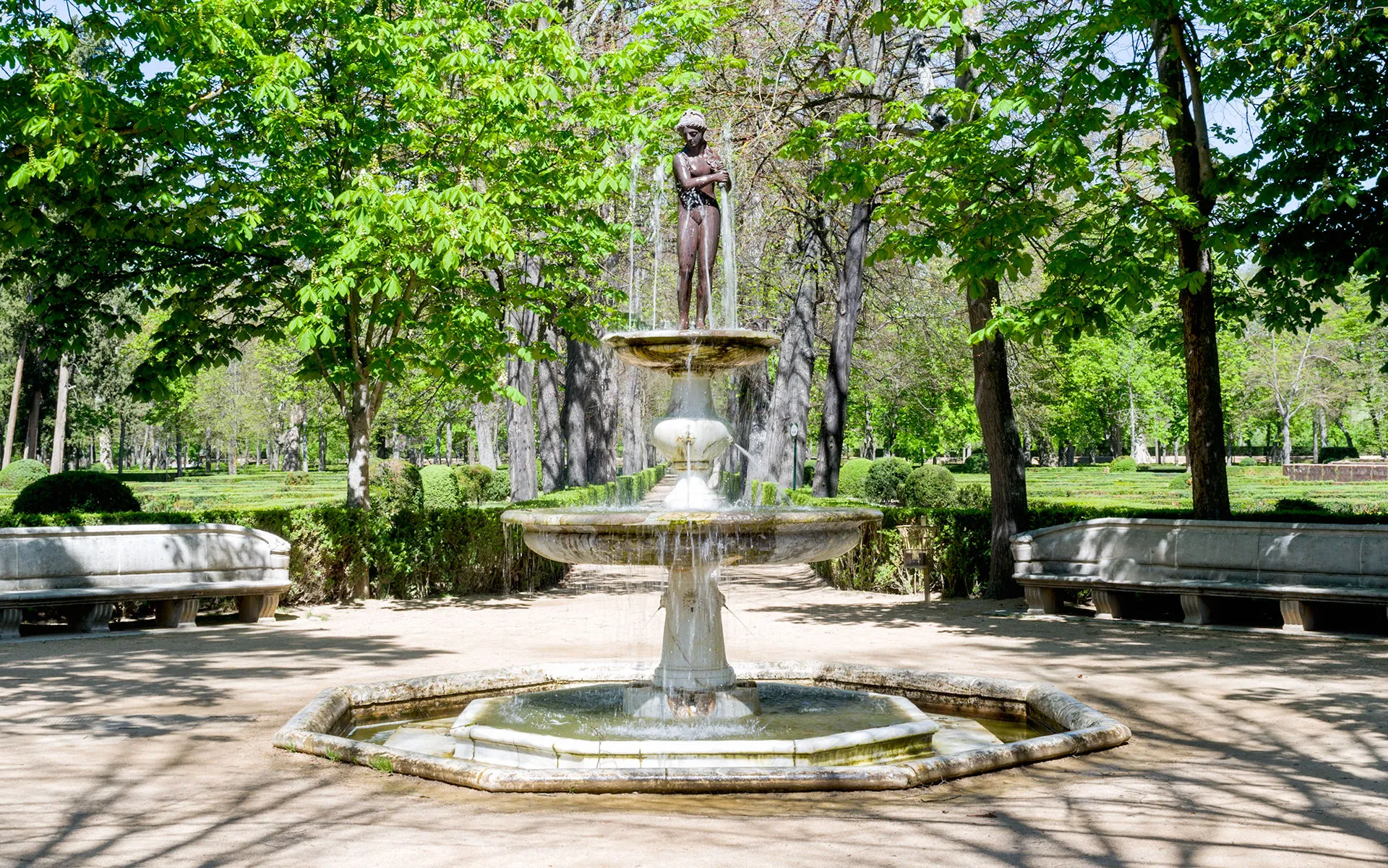 Fotografía de una fuente de mármol de dos tazas con figura de bronce en un parque verde.