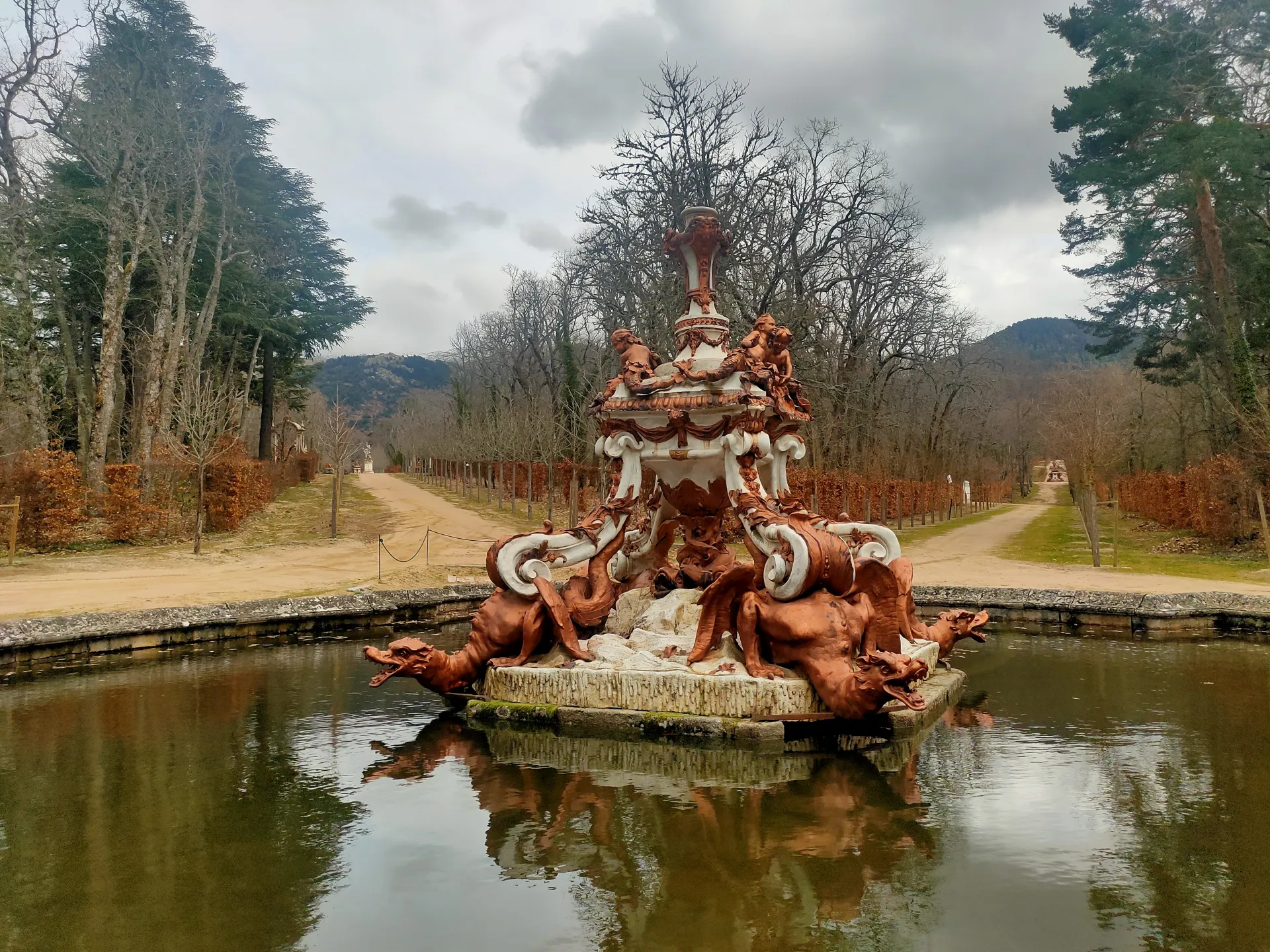 Fuente con remate en forma de jarrón sobre un pedestal decorado con dragones alados, en el centro de un estanque flanqueado por una alameda.