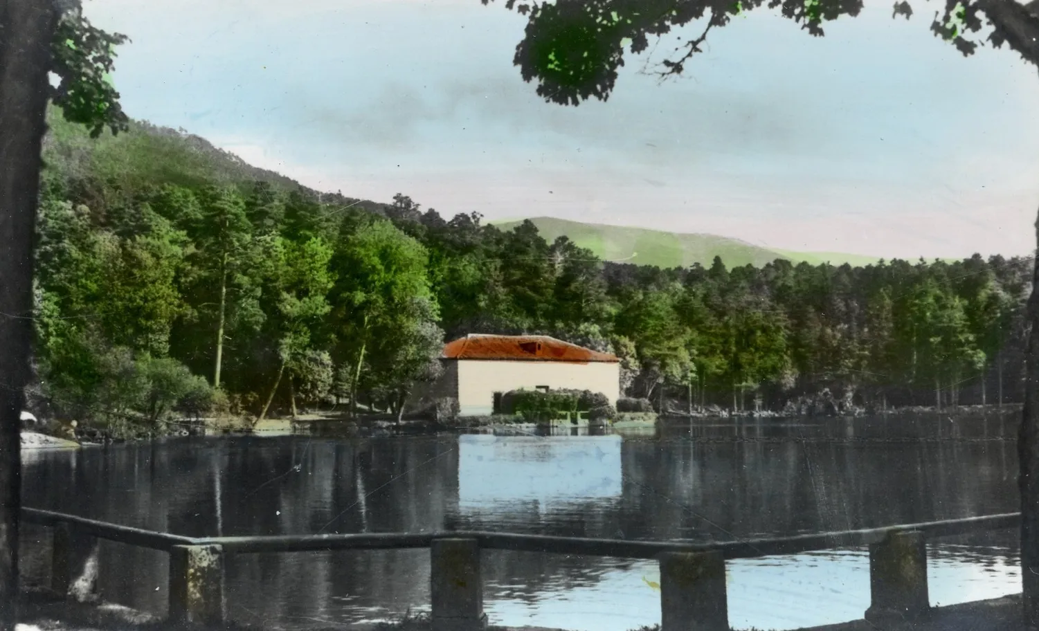 Fotografía antigua coloreada a mano de un estanque con una casa de tejado rojo entre árboles, montañas al fondo y valla de madera en primer plano.