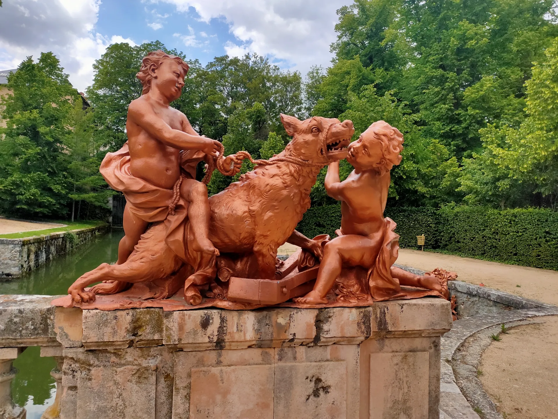 Grupo escultórico en terracota de dos niños sujetando con una cuerda a un lobo, sobre un pedestal de piedra al borde de un canal.