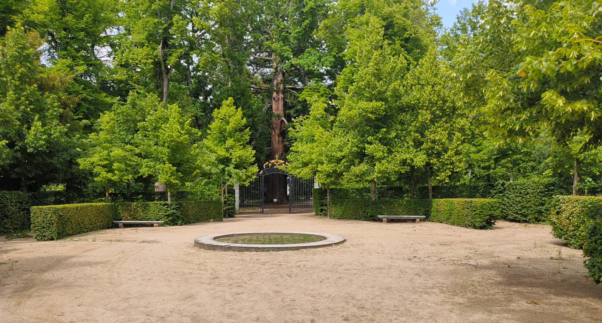 Explanada circular de tierra con un parterre bajo de piedra, bancos entre setos y una verja dorada junto a un gran árbol al fondo.
