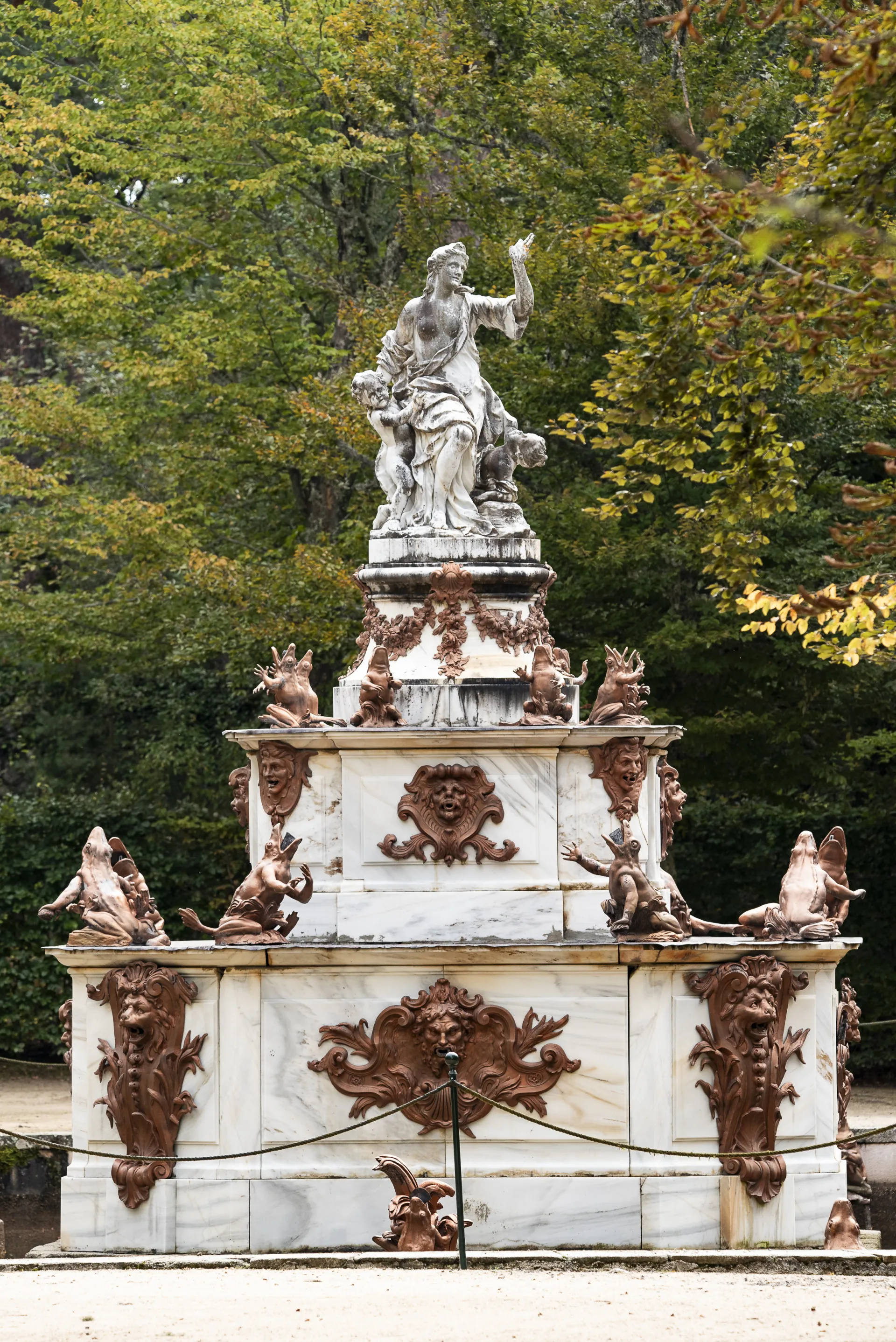 Escultura de una figura femenina con niños en lo alto de un pedestal de mármol escalonado, rodeado de ranas de bronce y máscaras decorativas.