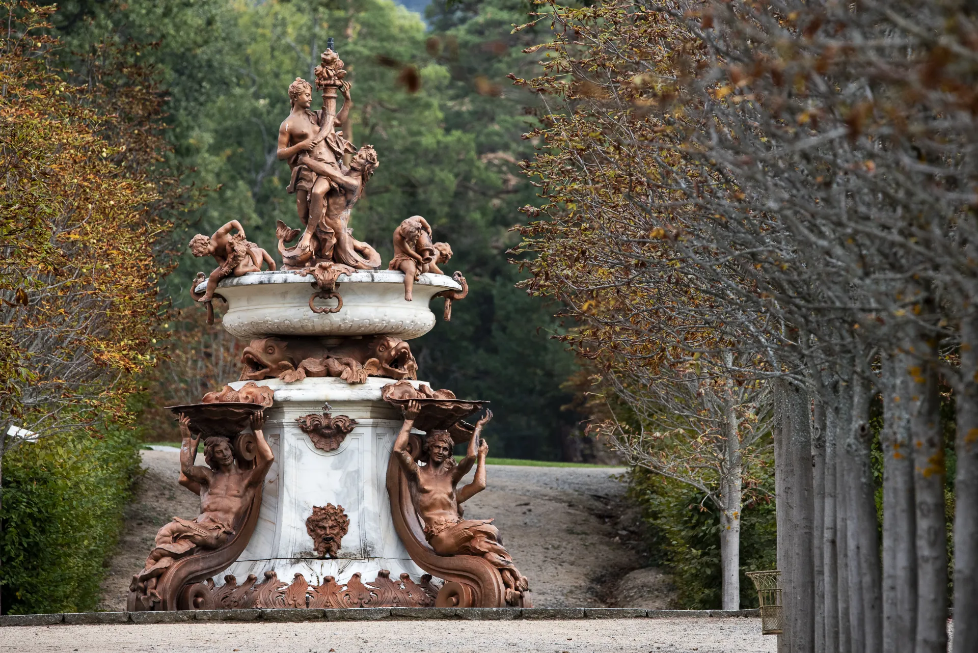 Fotografía de una fuente de mármol blanco y bronce con figuras y atlantes sosteniendo conchas, en un entorno boscoso otoñal.
