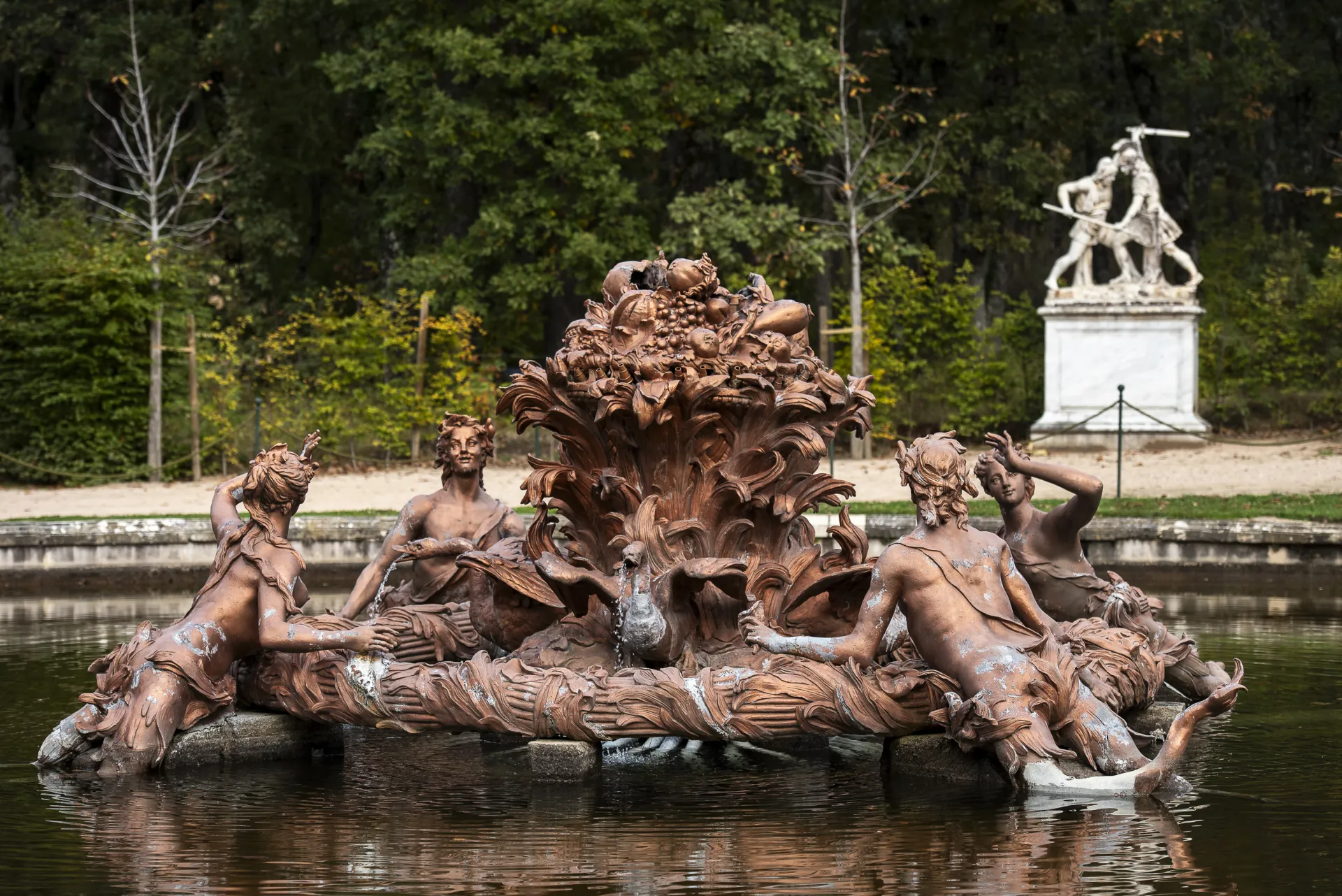 Escultura de bronce con figuras reclinadas rodeando un cisne surtidor, coronada por un canastillo de frutas, en una fuente rodeada de arbolado.