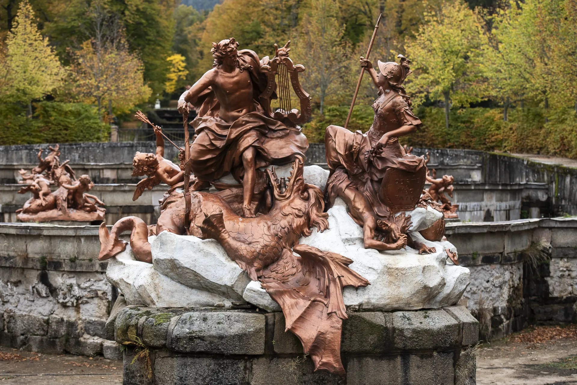 Fotografía de un grupo escultórico de bronce con figuras mitológicas sobre una roca, junto a una balaustrada de piedra y árboles con hojas otoñales.