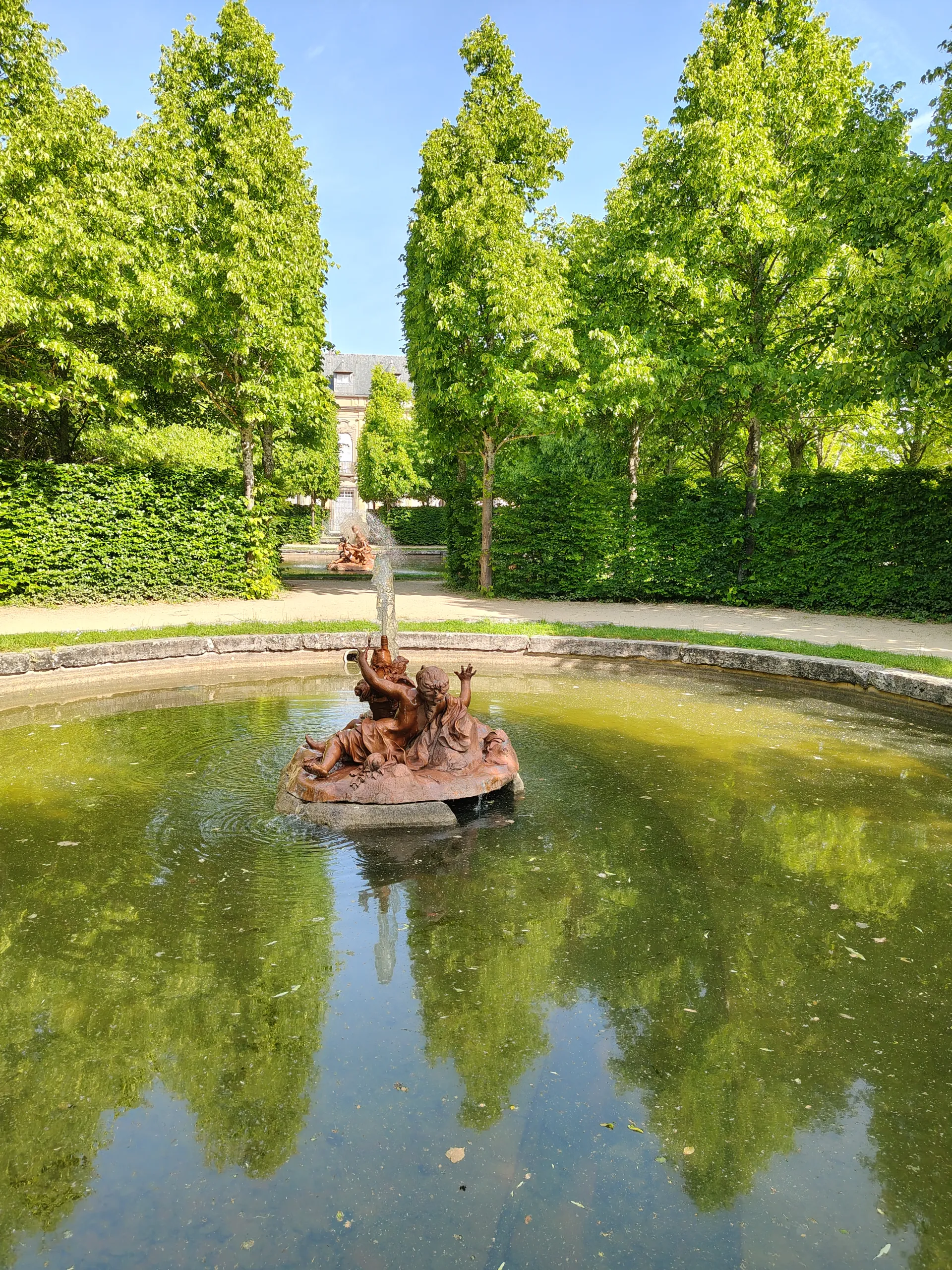Fotografía de una fuente con figuras de bronce sobre una base rocosa en un estanque, rodeada de árboles, con otra fuente y el palacio al fondo.