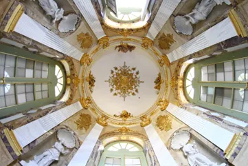 Fotografía tomada desde abajo de una cúpula interior decorada con estucos dorados y motivos barrocos, rodeada de ventanales.