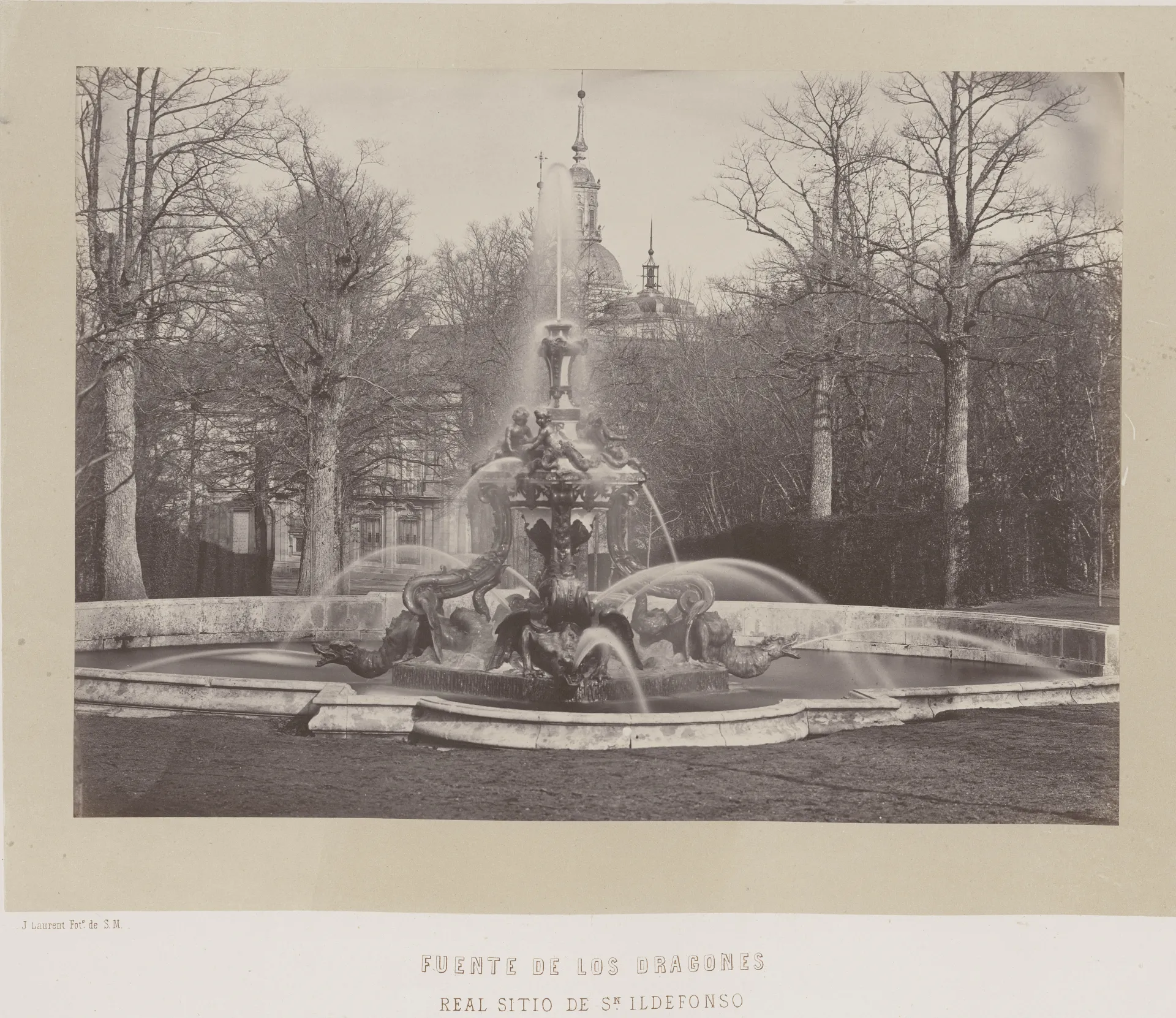 Fotografía antigua en blanco y negro de una fuente con surtidor vertical, entre árboles desnudos y la cúpula del palacio al fondo.