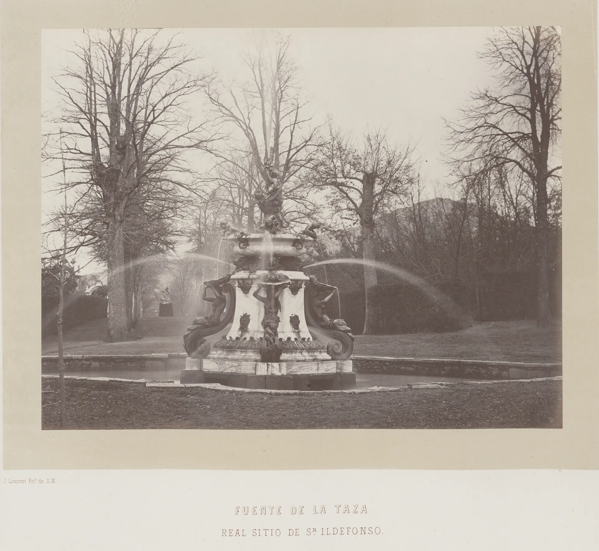 Fotografía histórica en blanco y negro de una fuente de piedra escalonada con chorros de agua en arco, entre árboles sin hojas.