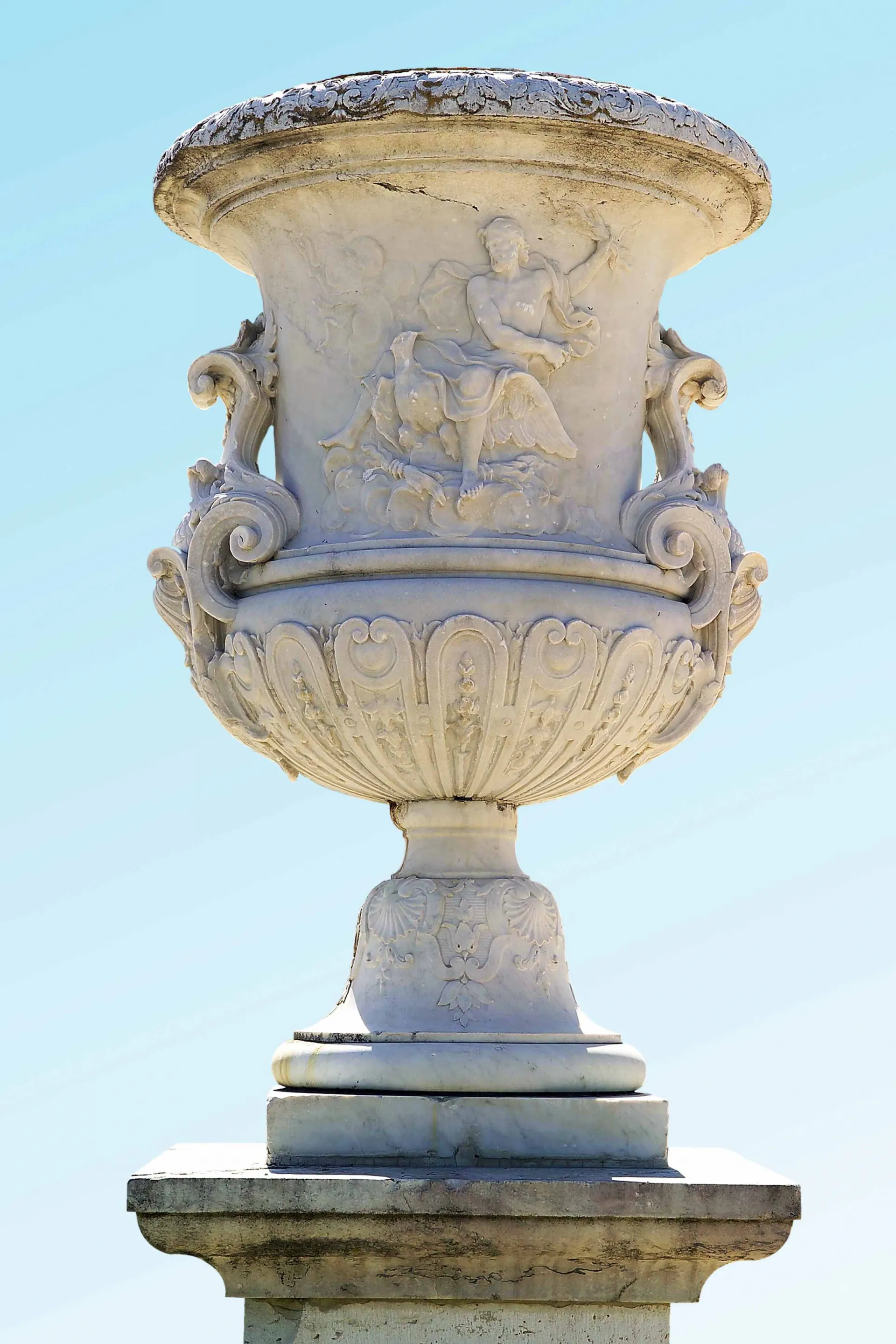 Fotografía de un jarrón de mármol con relieve de una figura clásica, sobre pedestal, contra un cielo despejado.