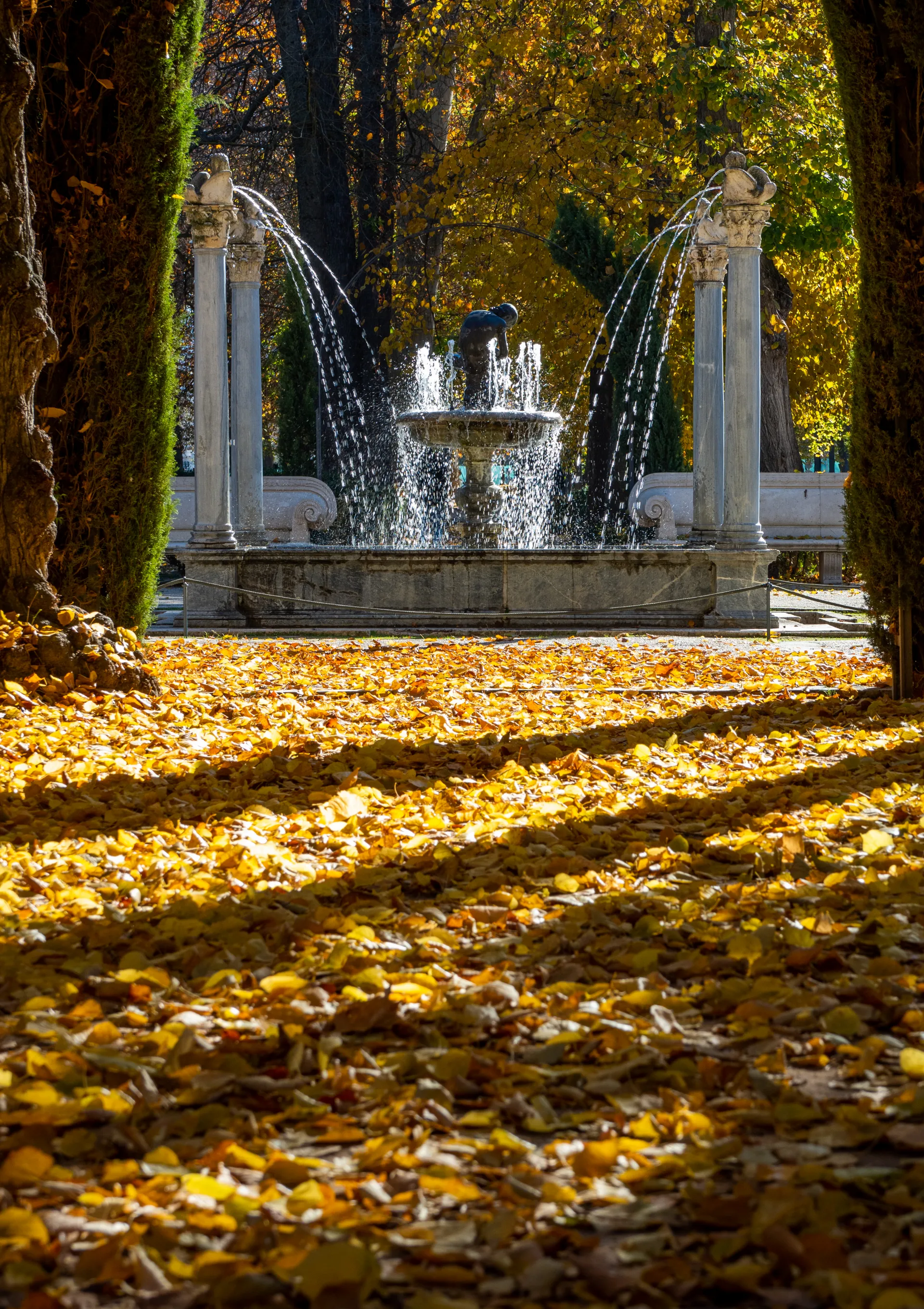 Fuente de las Arpías (o del Espinario) en Aranjuez, con columnas y surtidores de agua, en un paseo con hojarasca otoñal dorada.