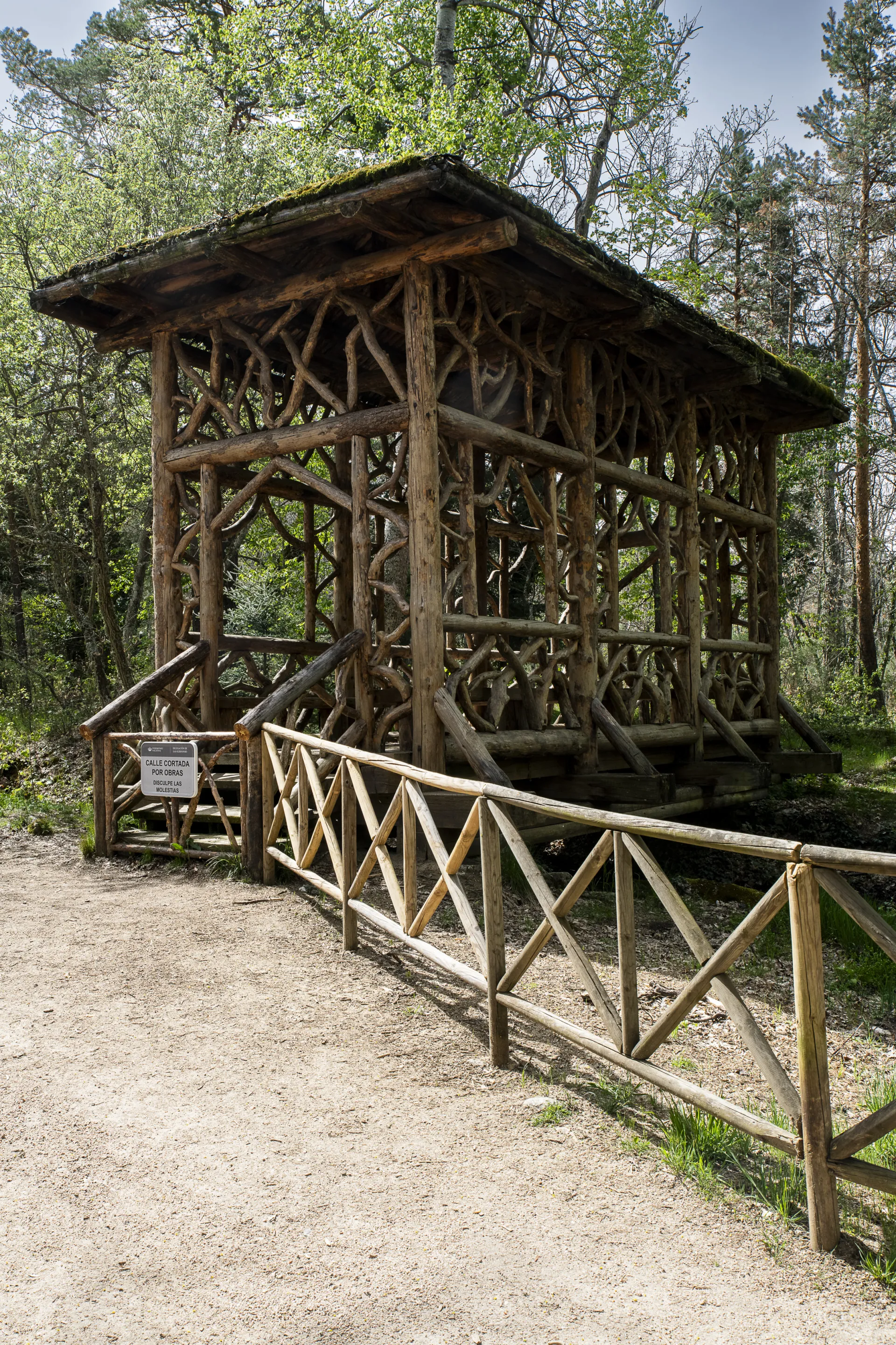 Pabellón-puente rústico construido con ramas y troncos retorcidos y tejado musgoso, en un bosque, con barandilla de madera en zigzag.