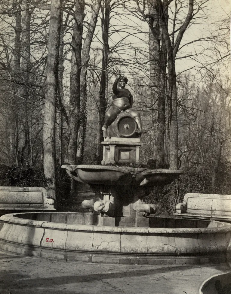 Fotografía antigua de la fuente de Baco con figura de bronce sobre un tonel, entre árboles.
