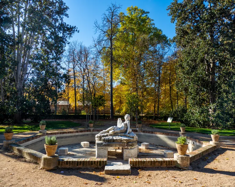 Escultura yacente de mármol en una fuente hundida del jardín, rodeada de arbolado otoñal.