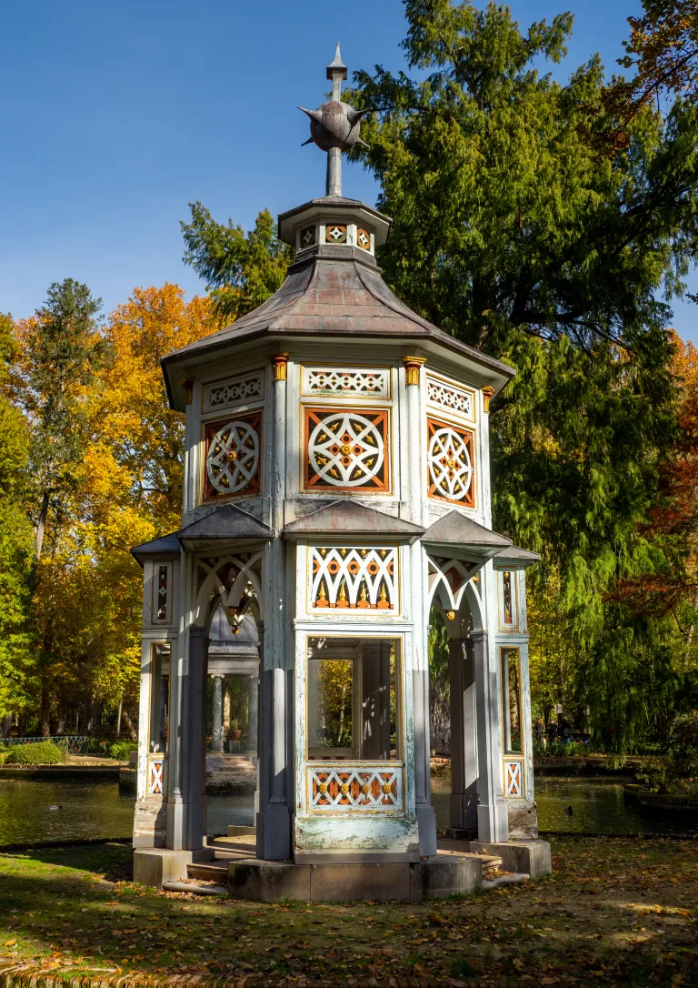 Fotografía de un templete chinesco blanco junto a un estanque, con arbolado otoñal.