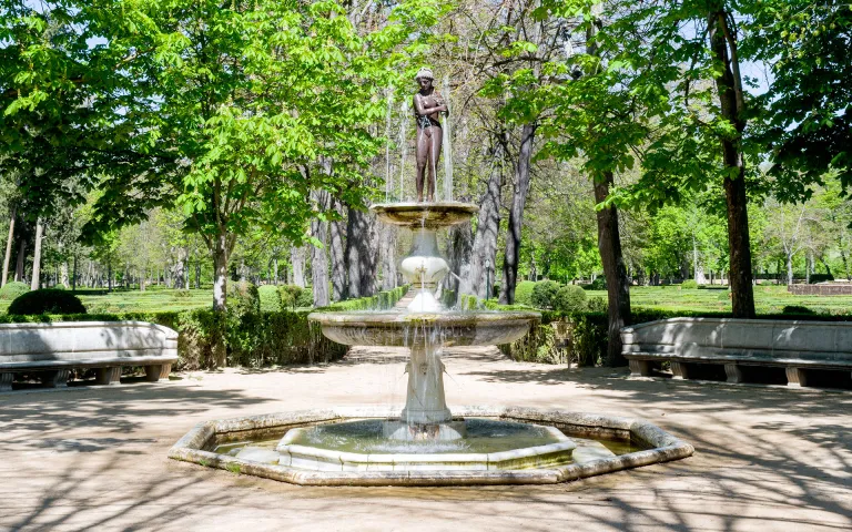 Fotografía de una fuente de mármol de dos tazas con figura de bronce en un parque verde.