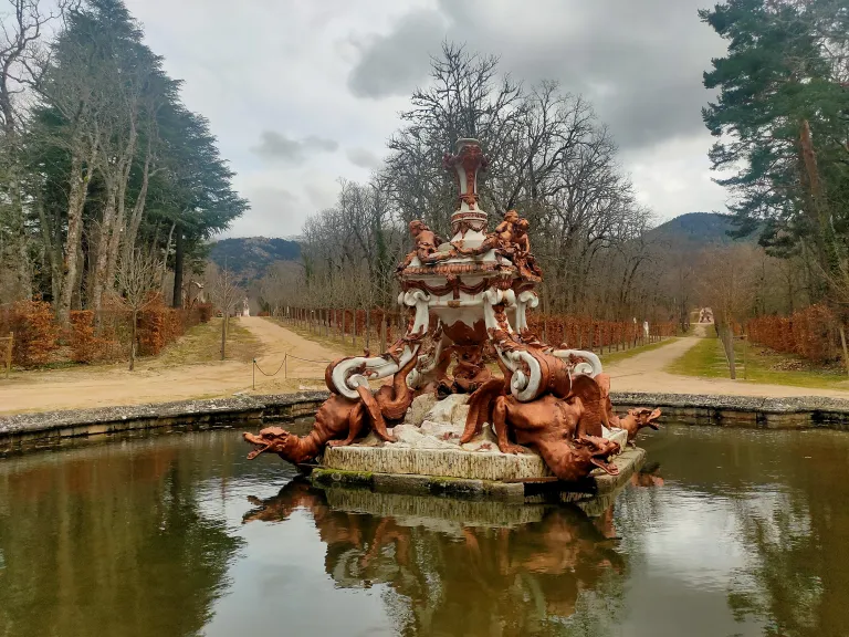 Fuente con remate en forma de jarrón sobre un pedestal decorado con dragones alados, en el centro de un estanque flanqueado por una alameda.
