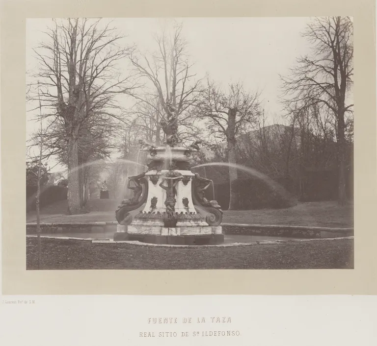 Fotografía antigua en blanco y negro de una fuente de dos tazas escalonadas con figuras en la base, entre árboles deshojados.