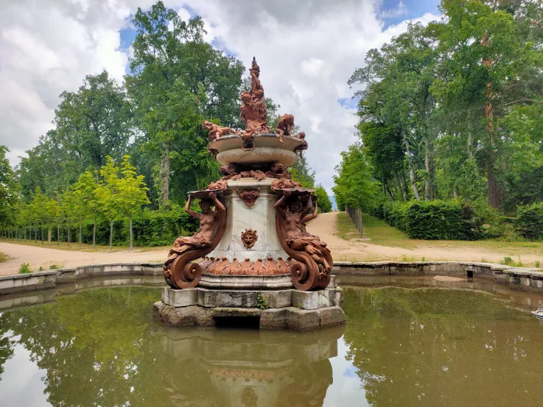 Fuente de dos tazas con figuras femeninas sosteniendo conchas y una máscara en el pedestal central, en un estanque junto a un camino arbolado.