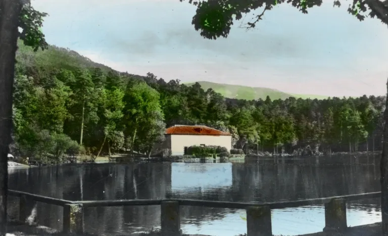 Fotografía antigua coloreada a mano de un estanque con una casa de tejado rojo entre árboles, montañas al fondo y valla de madera en primer plano.