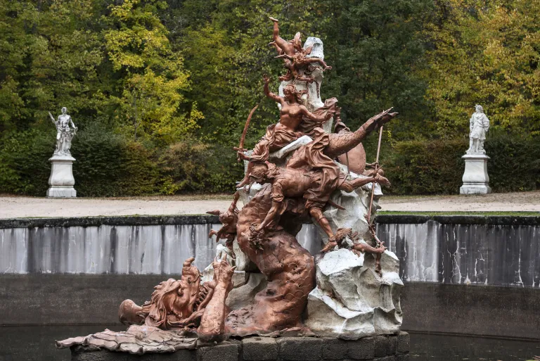 Grupo escultórico de bronce con figuras mitológicas sobre roca y un monstruo marino, en el centro de una fuente flanqueada por estatuas de piedra.
