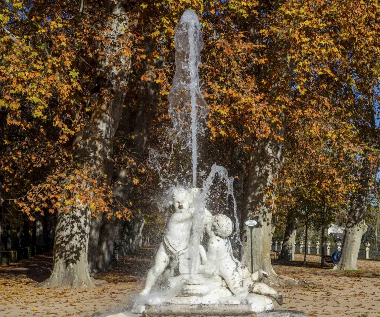 Fuente de la Boticaria con el surtidor de agua en marcha, rodeada de árboles con follaje otoñal anaranjado.