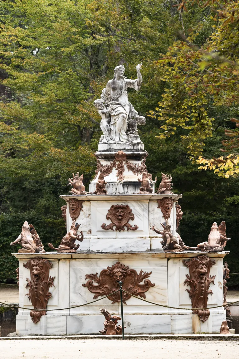 Escultura de una figura femenina con niños en lo alto de un pedestal de mármol escalonado, rodeado de ranas de bronce y máscaras decorativas.