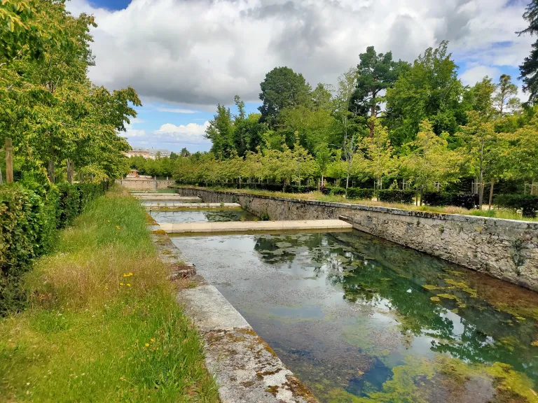 Fotografía de un canal de agua entre muros de piedra y arboledas, con el palacio al fondo entre nubes.