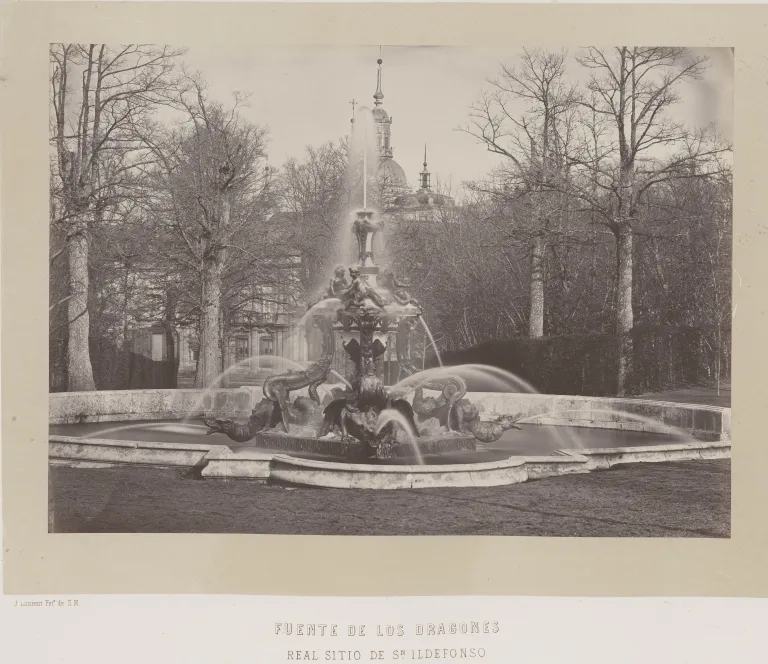 Fotografía antigua en blanco y negro de una fuente con surtidor vertical, entre árboles desnudos y la cúpula del palacio al fondo.