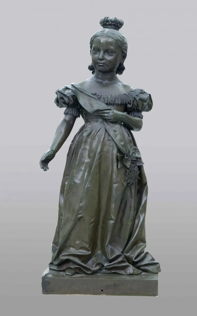 Escultura de bronce de una niña con vestido de gala, banda cruzada con una condecoración y corona sobre el pelo recogido.