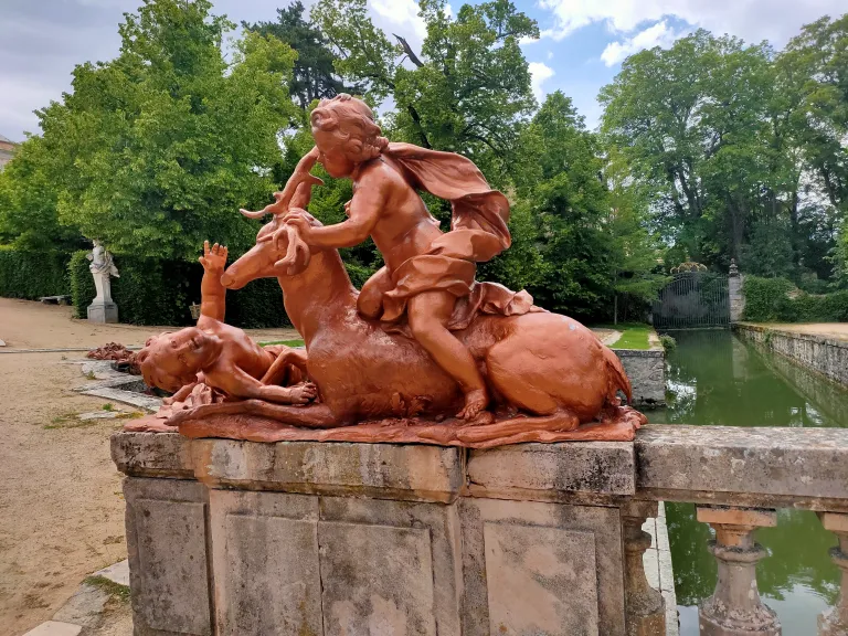 Escultura de terracota de un niño sujetando por la cornamenta a un ciervo, con otro niño tumbado junto a ellos, sobre un pretil de piedra junto a un estanque.