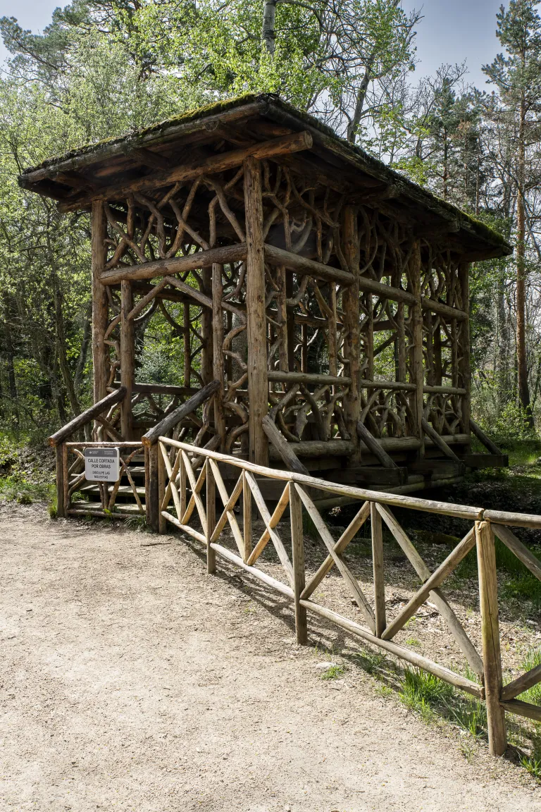 Pabellón-puente rústico construido con ramas y troncos retorcidos y tejado musgoso, en un bosque, con barandilla de madera en zigzag.